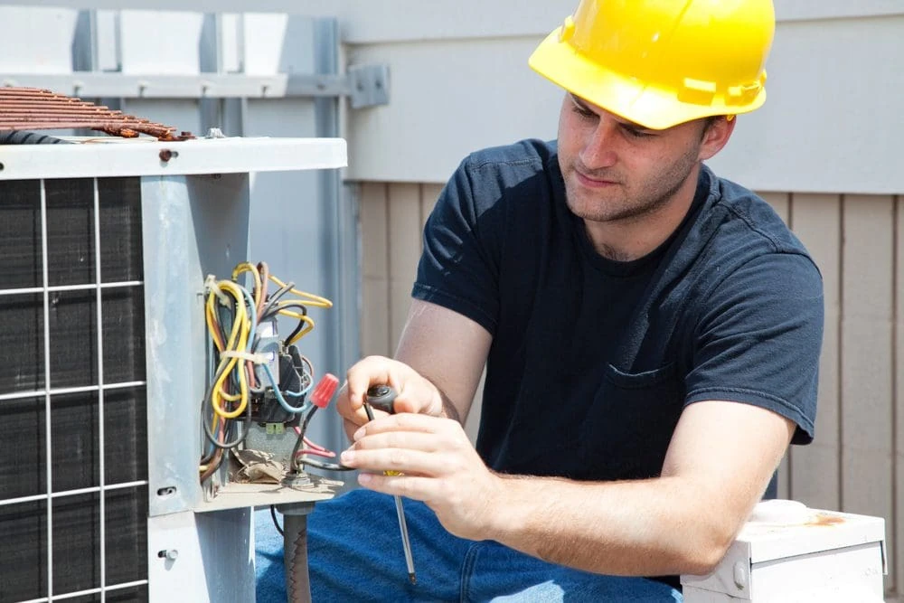 Technician in a yellow hard hat checking wiring inside an outdoor HVAC unit